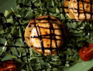 Nahaufname des appetitlich angerichteter Tellers mit gegrilltem Käsetaler mit Grillmuster auf einem Bett aus Rucola, garniert mit halbierten Kirschtomaten und einer dunklen Balsamico-Glasur im Restaurant Wulfsmühle.