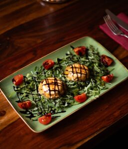 Ein appetitlich angerichteter Teller auf einem Holztisch: Zwei runde, gegrillte Käsetaler mit Grillmuster auf einem Bett aus Rucola, garniert mit halbierten Kirschtomaten und einer dunklen Balsamico-Glasur. Vom Restaurant Wulfsmühle.