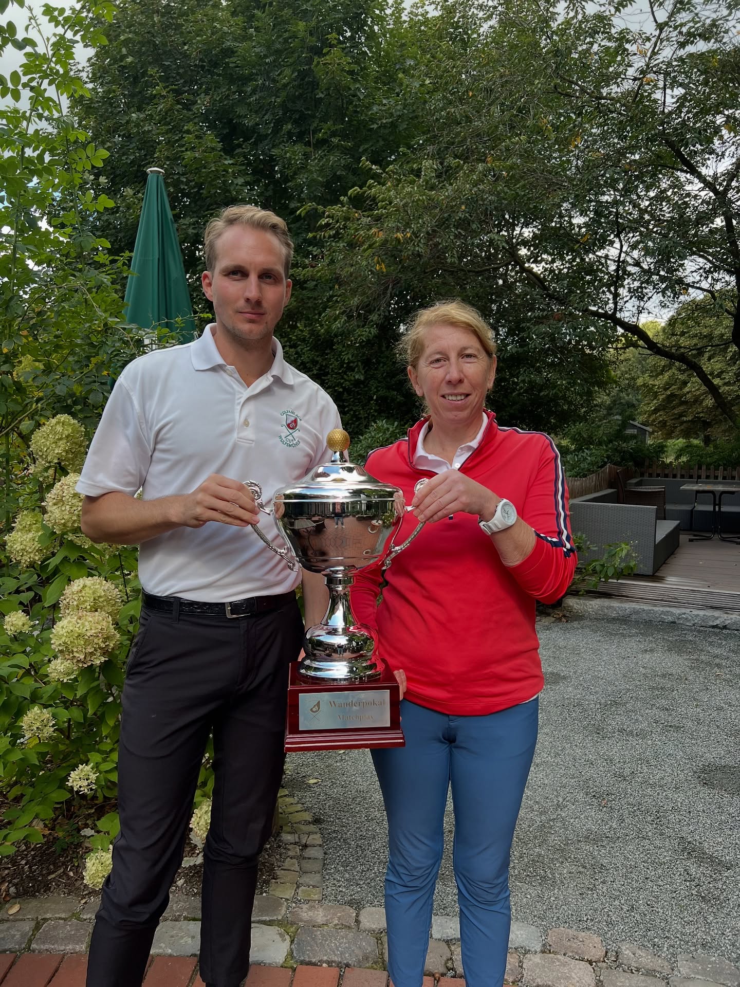 Ein Mann und eine Frau stehen im Freien auf einer Golfanlage und halten gemeinsam einen großen silbernen Pokal.