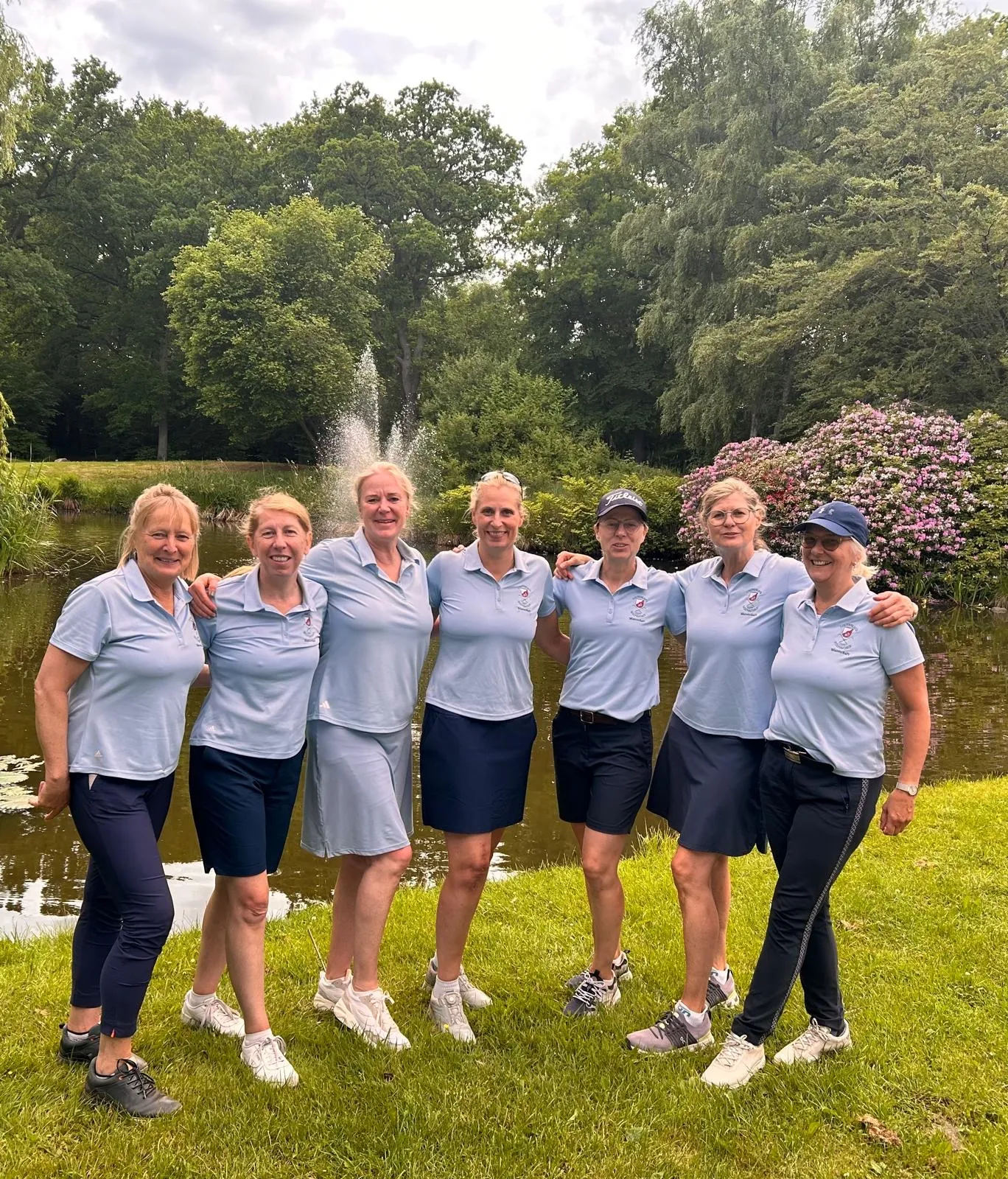 Sieben Frauen in hellblauen Team-Poloshirts posieren vor einem Teich mit Springbrunnen