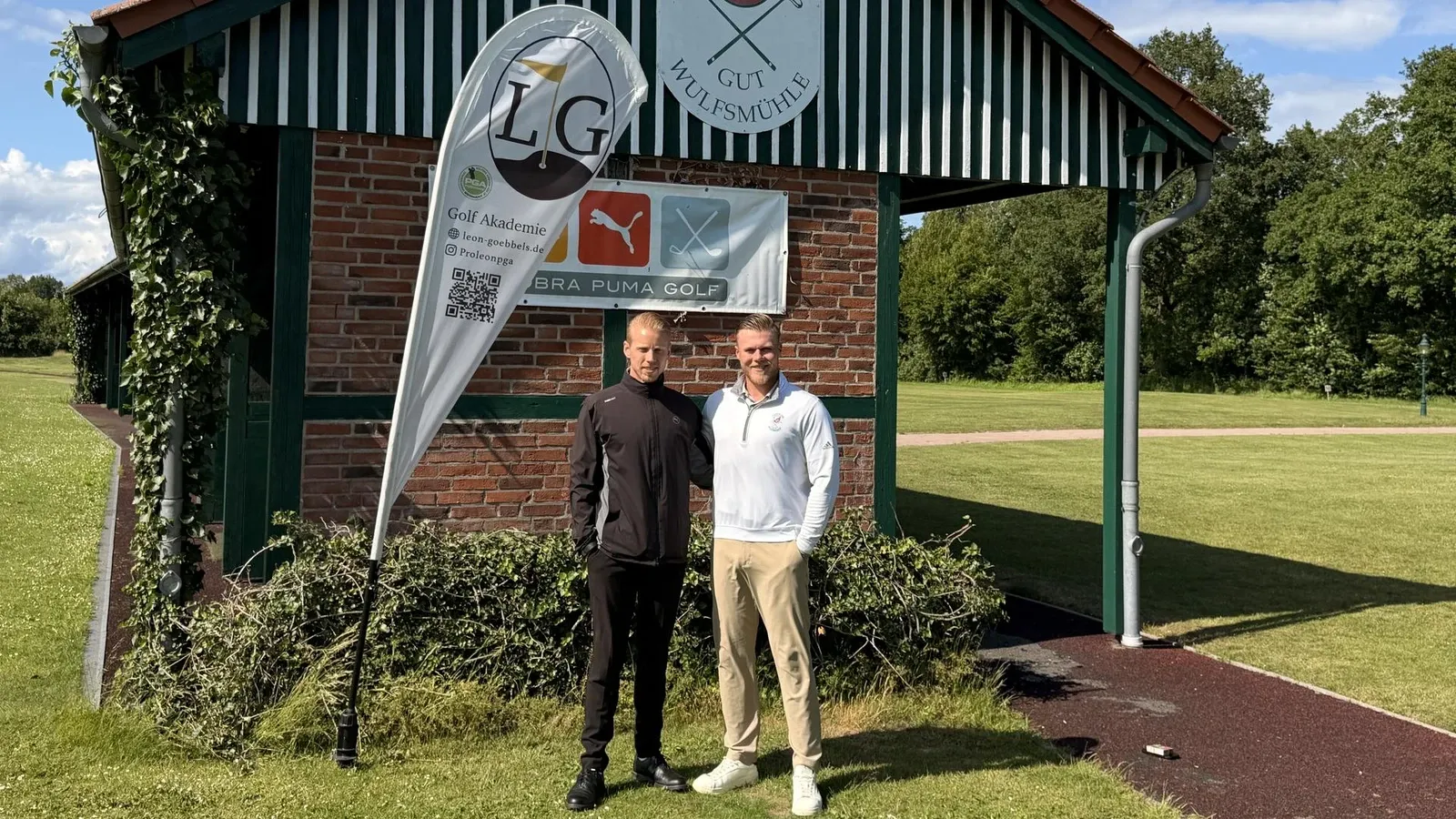 Zwei Männer vor dem Gebäude der Golf Akademie Gut Wulfsmühle mit einer LG Golf Akademie Fahne
